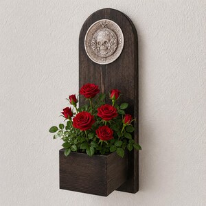 May include: A dark wooden wall planter with a carved skull design and a rectangular planter box. The planter holds vibrant red roses with green leaves. The skull design is on a circular plaque.