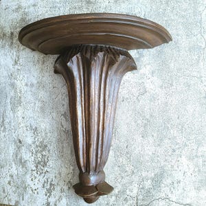 May include: A dark brown, carved wooden wall shelf. The shelf has a rounded top and a decorative, fluted support that resembles a stylized flower. The shelf is designed to be mounted on a wall and is made of wood.
