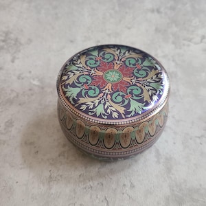 May include: A round, decorative tin with a detailed floral design on the lid. The lid features a central red flower surrounded by green and gold foliage against a dark blue background. The sides have a repeating pattern.