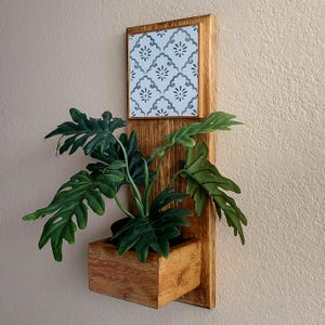 May include: A wall-mounted wooden planter featuring a rectangular backboard with a square tile insert at the top. The tile displays a repeating gray floral pattern on a white background. A green philodendron plant sits inside the wooden box base.