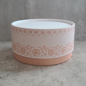 May include: A round, two-piece gift box with a white lid and a peach-colored base. The box's body is decorated with a delicate, peach-colored lace pattern. The box is designed for gifting and storage.