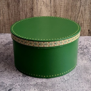 May include: A round, green box with a lid. The box has a decorative band around the middle with a pattern of red and green leaves. The lid and base are trimmed with small, gold-colored dots. The box is sitting on a gray surface.