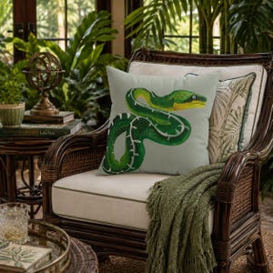 May include: A square throw pillow featuring a detailed illustration of a coiled green snake with yellow accents on a light gray background. The snake has a visible eye and white markings along its body, centered on the pillow's front surface.