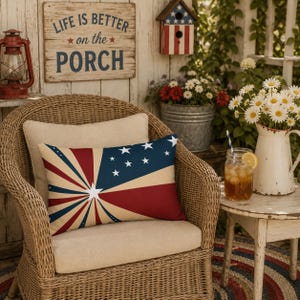 May include: A rectangular throw pillow featuring a patriotic design with a blue corner containing white stars and red, white, and blue sunburst rays radiating from a central white star, resting on a woven wicker chair with a beige seat cushion.