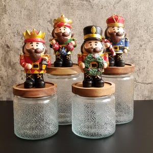 May include: Set of four glass jars with wooden lids, each topped with a decorative nutcracker figurine. The nutcrackers wear colorful uniforms and hats, holding various festive items. The jars have a patterned design.