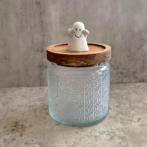 May include: A clear glass jar with a geometric pattern, topped with a wooden lid and a small white ceramic ghost figurine. The ghost has a smiling face. The jar is likely for storage or decoration.