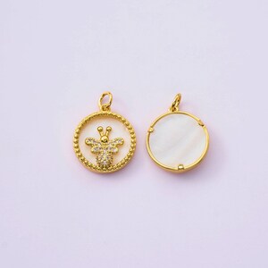 May include: Two gold-colored pendants. One pendant features a bee design with clear crystals on a white background, surrounded by a beaded gold border. The other pendant has a white, circular mother-of-pearl design with a gold border.