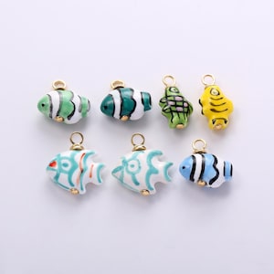 May include: A collection of seven small, colorful fish-shaped charms, each featuring a gold-tone metal jump ring at the top. The charms display various patterns and colors, including green, white, yellow, blue, and turquoise, with painted details.