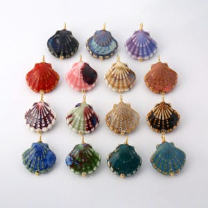 May include: A collection of sixteen ceramic seashell pendants arranged in a grid. Each pendant features a unique glaze color, including blue, red, pink, brown, green, and purple, with a small gold-toned metal loop attached to the top for hanging.