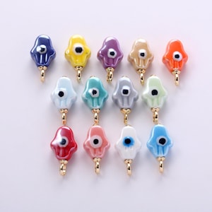 May include: A collection of fourteen small hamsa hand charms in various glossy colors including blue, yellow, purple, beige, orange, light blue, teal, gray, green, red, pink, and white, each featuring a central eye detail and a gold metal loop.