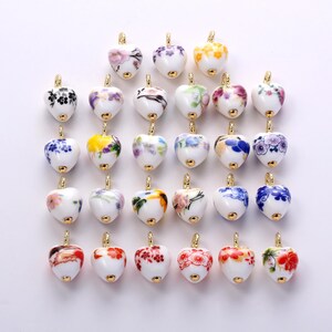 May include: A collection of twenty-four heart-shaped ceramic charms, each featuring a unique hand-painted floral pattern in various colors. Every charm is finished with a small gold-toned metal loop at the top for attachment to jewelry.
