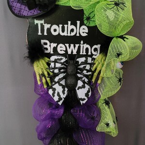 May include: A Halloween-themed decoration with a witch hat, green mesh, and a sign that reads "Trouble Brewing." The decoration features a black spider, purple mesh, and a pair of purple witch boots. Spiders are scattered throughout the design.