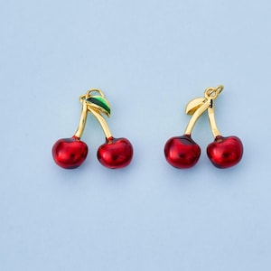 May include: Two cherry-shaped earrings with a glossy red finish. The stems and leaves are gold and green. The earrings are set against a light blue background.