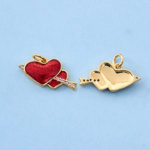 May include: Two gold-toned metal heart charms with an arrow motif. One charm features a red glitter enamel finish and small clear stones on the arrow, while the other is a plain polished gold-toned metal version with a smooth surface.