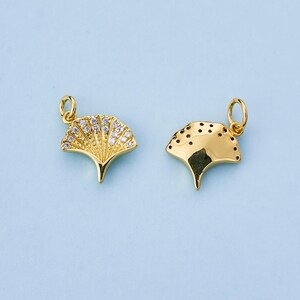 Op de afbeelding: Twee goudkleurige ginkgo blad bedels. Eén bedel is versierd met kleine, heldere kristallen, terwijl de andere een glad oppervlak heeft met kleine, gelijkmatig verdeelde gaatjes. Beide bedels hebben een kleine lus aan de bovenkant om te bevestigen.