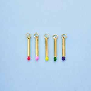 May include: Five gold-coloured matchstick charms with colourful tips: red, pink, yellow, green, and purple. Each charm has a small loop at the top for attaching to jewellery. The background is a light blue.
