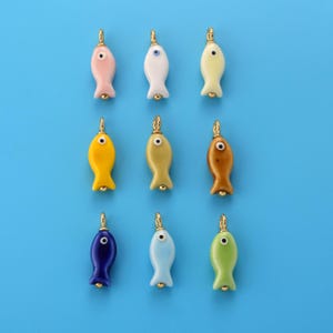 May include: A collection of nine small fish-shaped charms arranged in a grid. Each charm features a smooth, solid-coloured body in shades of pink, white, yellow, gold, brown, dark blue, light blue, and green, with a single eye and a gold metal loop.