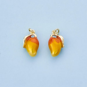 May include: Two gold-colored mango charms with yellow and orange enamel. Each charm has a gold-colored loop at the top and a gold-colored leaf detail. The charms are set against a light blue background.