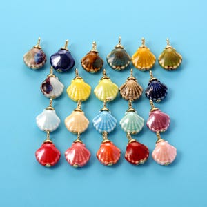 May include: A collection of small, glossy ceramic seashell charms in various colors including red, pink, blue, yellow, and brown. Each charm features a gold-toned metal loop at the top for attachment, showcasing a ribbed, fan-like shell design.