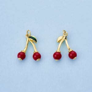 May include: Two gold-colored cherry earrings with red cherry beads and green and gold leaves. The earrings are set against a light blue background. The earrings are a fun and playful accessory.
