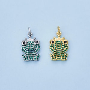 May include: Two frog-shaped pendants. One is silver-toned, the other gold-toned. Both feature a crown and are encrusted with green and black gemstones. The pendants are designed with a small loop at the top for hanging.