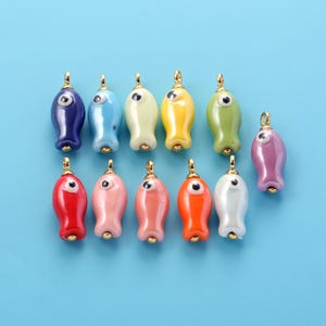 May include: A collection of eleven small fish-shaped charms made of smooth, glossy material in various colors including dark blue, light blue, yellow, green, red, pink, orange, and white, each featuring a single eye and a gold-toned metal loop.