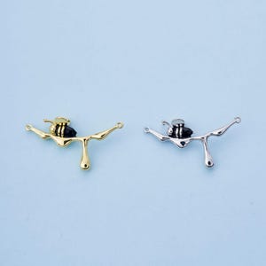 Könnte beinhalten: Zwei bienenförmige Anhänger, einer goldfarben und einer silberfarben, jeweils mit schwarzem Körper und tropfendem Honigdesign. Die Anhänger haben kleine Schlaufen an jedem Ende zum Befestigen.