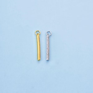 May include: Two bar-shaped pendants, one gold and the other silver, against a light blue backdrop. The gold pendant has a smooth surface, while the silver is encrusted with small, sparkling stones. Both have a small loop at the top for attachment.