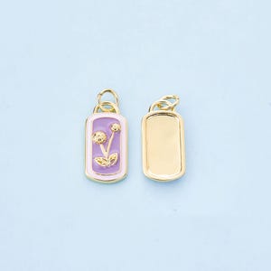 Puede incluir: Dos colgantes rectangulares dorados. Uno presenta un diseño floral morado con detalles dorados y un borde rosa claro. El otro colgante es un rectángulo dorado liso. Ambos colgantes tienen un pequeño lazo en la parte superior para sujetar.