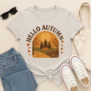 Può includere: Una maglietta colorata con la scritta "HELLO AUTUMN" ad arco sopra un disegno di paesaggio autunnale. Il disegno presenta un sole, alberi e foglie che cadono in tonalità marroni e arancioni. La maglietta è abbinata a jeans blu e scarpe da ginnastica bianche.