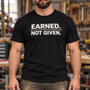 Può includere: Una t-shirt nera a maniche corte con girocollo, caratterizzata dalla scritta bianca EARNED. NOT GIVEN. stampata in un carattere sans-serif in grassetto sul petto, con una linea orizzontale che separa le due frasi del design.