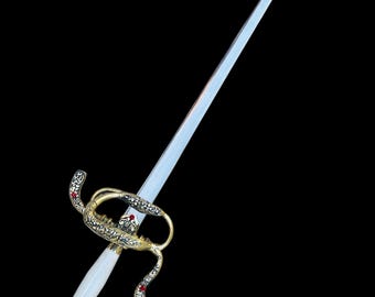 May include: A rapier with a long, slender, double-edged steel blade. The hilt features an ornate gold-toned metal guard with intricate filigree patterns and small red stone accents. The handle is smooth and white, ending in a decorative pommel.