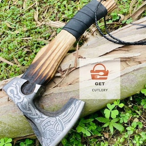 May include: A handcrafted axe with a wooden handle and a metal blade. The handle is wrapped in black leather. The blade features intricate engraved designs. The axe is displayed on a log with green foliage in the background. The text "GET CUTLERY" is visible.