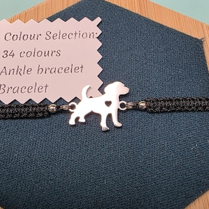 May include: A black macrame bracelet featuring a silver dog charm with a heart detail. The bracelet is presented on a blue fabric and a wooden base. A tag reads "Colour Selection: 34 colours Ankle bracelet Bracelet."