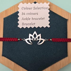 Puede incluir: Una pulsera con dije de flor de loto plateada con un cordón tejido rojo oscuro. La pulsera se exhibe sobre un fondo verde azulado oscuro, con una etiqueta que indica 34 opciones de color y los términos "Tobillera" y "Pulsera".