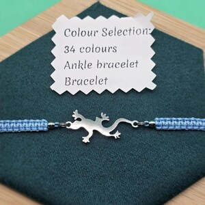 May include: A light blue macrame ankle bracelet with a silver lizard charm. The bracelet is displayed on a dark teal surface with a wooden frame. A white tag indicates color options.
