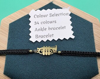 May include: A black macrame bracelet with a gold-colored trumpet charm. A white tag displays "Colour Selection: 34 colours Ankle bracelet Bracelet". The bracelet is displayed on a teal fabric and a wooden surface.