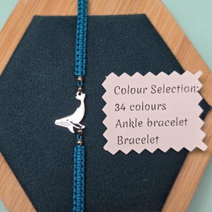 Può includere: Un braccialetto in macramè turchese con un ciondolo a forma di balena argentata. Il braccialetto è esposto su una superficie esagonale blu scuro. Un'etichetta indica una selezione di 34 colori, cavigliera o braccialetto.