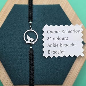 Pode incluir: Pulseira preta trançada com um pingente de lobo prateado. O pingente apresenta um lobo uivando dentro de um círculo. A imagem inclui uma etiqueta com "Colour Selection: 34 colours, Ankle bracelet, Bracelet."