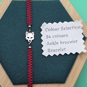 Puede incluir: Una pulsera de macramé burdeos con un dije de cabeza de lobo plateado. La pulsera se muestra sobre una superficie hexagonal verde oscuro. La imagen incluye una etiqueta que dice "Colour Selection: 34 colours Ankle bracelet Bracelet."