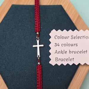 Peut inclure: Un bracelet en macramé bordeaux avec un charm en forme de croix argentée. Le bracelet est présenté sur une surface bleu canard foncé dans un cadre en bois. Une étiquette indique un choix de 34 couleurs.