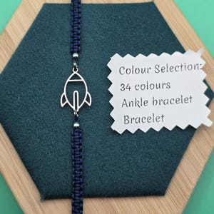 May include: A navy blue macrame bracelet with a silver rocket charm. The bracelet is displayed on a dark green hexagonal surface. The text on the card reads: Colour Selection: 34 colours, Ankle bracelet, Bracelet.