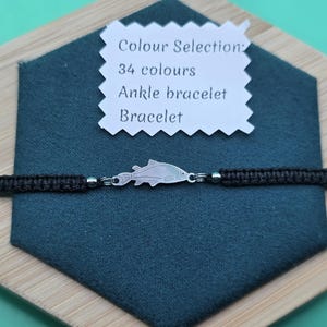 May include: Black ankle bracelet with a silver fish charm. The bracelet is displayed on a dark teal surface, with a white tag that reads "Colour Selection: 34 colours Ankle bracelet Bracelet."