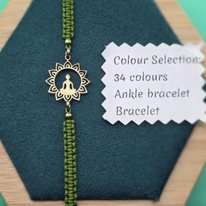 May include: A green macrame bracelet with a gold-colored charm featuring a seated Buddha figure within a sunburst design. The image includes a tag with text: "Colour Selection: 34 colours Ankle bracelet Bracelet."
