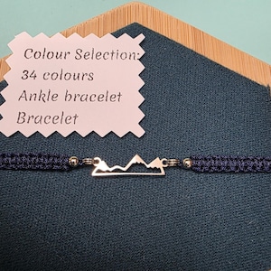 May include: A navy blue macrame bracelet with a silver mountain range charm. The bracelet is displayed on a teal background with a wooden accent. A tag reads "Colour Selection: 34 colours, Ankle bracelet, Bracelet."