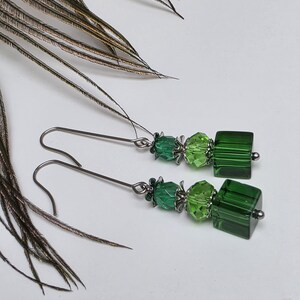 Può includere: Un paio di orecchini pendenti con perline di vetro verde e cristalli quadrati color smeraldo. Gli orecchini hanno ganci per le orecchie color argento e dettagli metallici decorativi. Il design include varie tonalità di verde, creando un accessorio vivace.