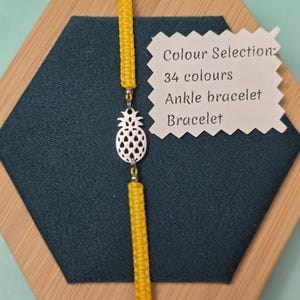 May include: A yellow ankle bracelet with a silver pineapple charm. The bracelet is displayed on a dark teal surface with a white tag that reads "Colour Selection: 34 colours Ankle bracelet Bracelet."