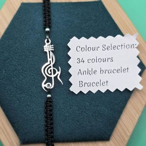 Op de afbeelding: Een zwarte macramé enkelband met een zilverkleurige vioolsleutel bedel. De armband wordt gepresenteerd op een donkerblauwe ondergrond, met een wit label met de tekst "Colour Selection: 34 colours Ankle bracelet Bracelet."