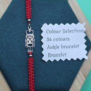 Puede incluir: Una pulsera de macramé roja con un dije de cascanueces plateado. La pulsera se exhibe sobre una superficie hexagonal verde oscuro. Una etiqueta blanca indica opciones de color y el estilo de la pulsera.