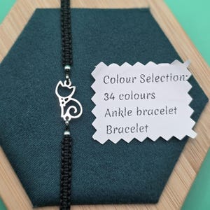 May include: A black macrame ankle bracelet with a silver cat charm. The charm is a stylized cat silhouette. The bracelet is displayed on a dark green hexagonal surface with a wooden border. Text on a white tag reads "Colour Selection: 34 colours, Ankle bracelet, Bracelet."